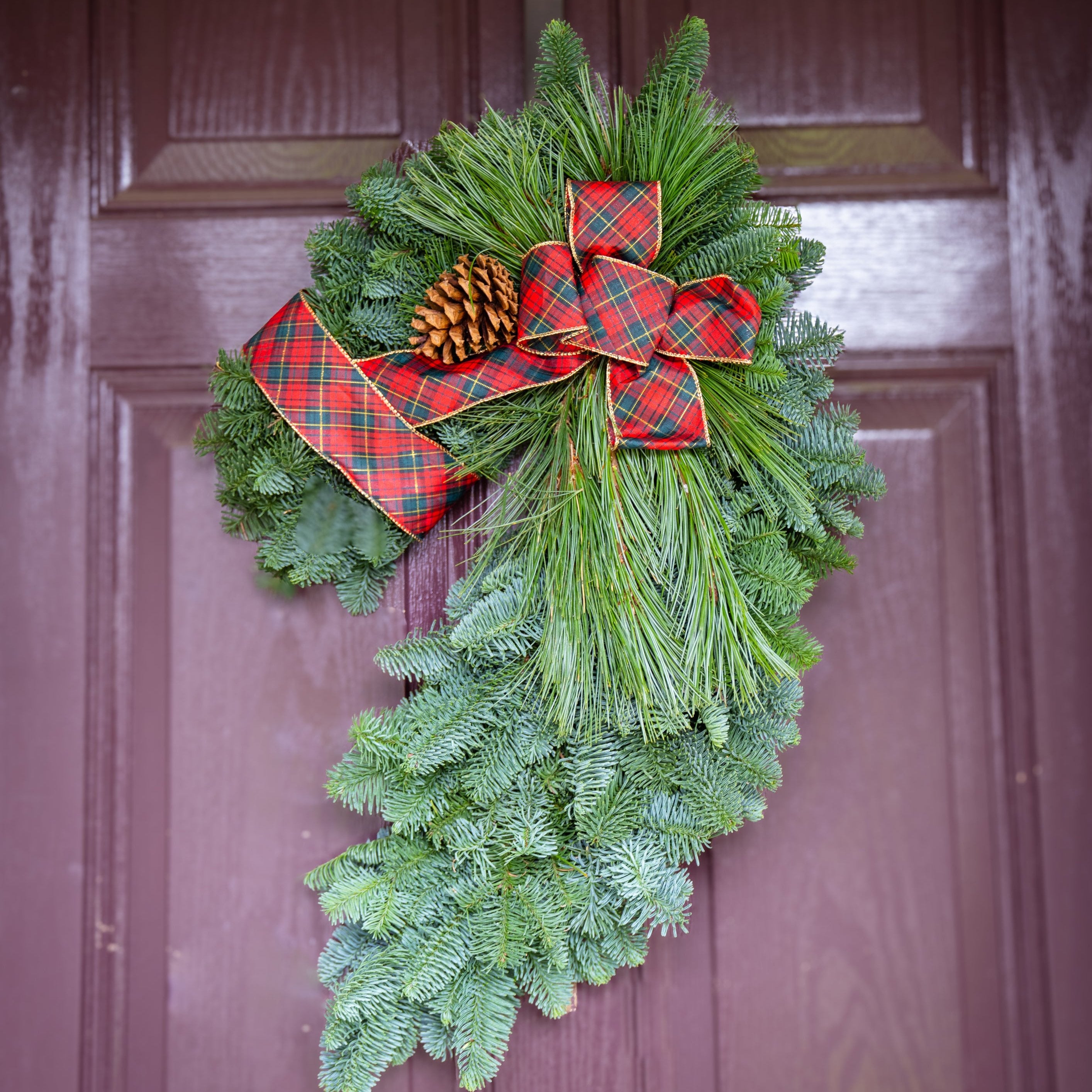 A handcrafted holiday wreath in the shape of a horse's head, made with green noble fir and adorned with pine cones, white pine, and a red bow.
