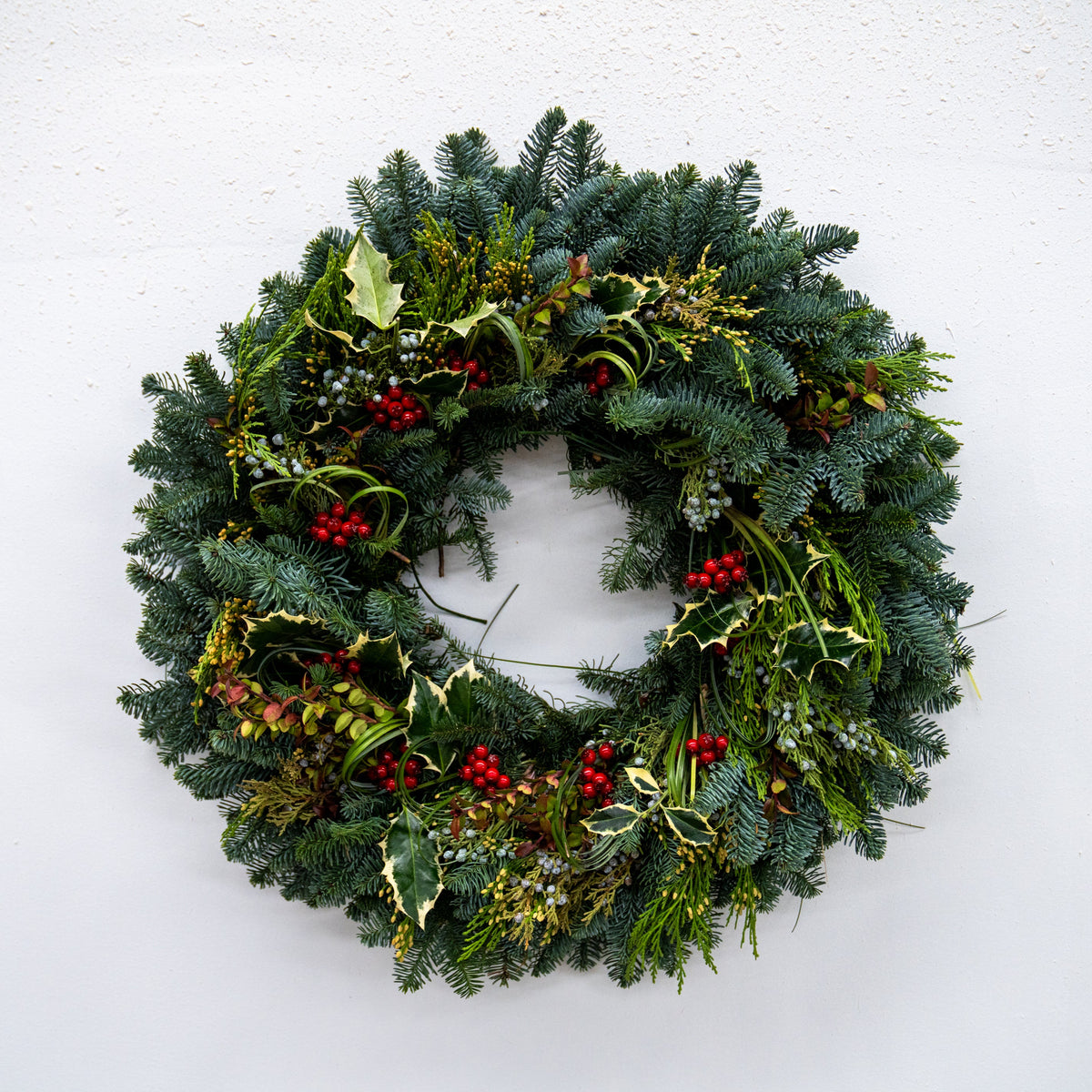 A festive Christmas wreath made of green pine and fir branches, decorated with small red berries, holly leaves, and gold mistletoe, hanging against a plain white wall.