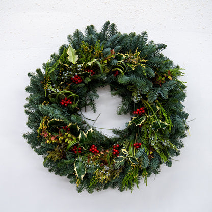 A festive Christmas wreath made of green pine and fir branches, decorated with small red berries, holly leaves, and gold mistletoe, hanging against a plain white wall.